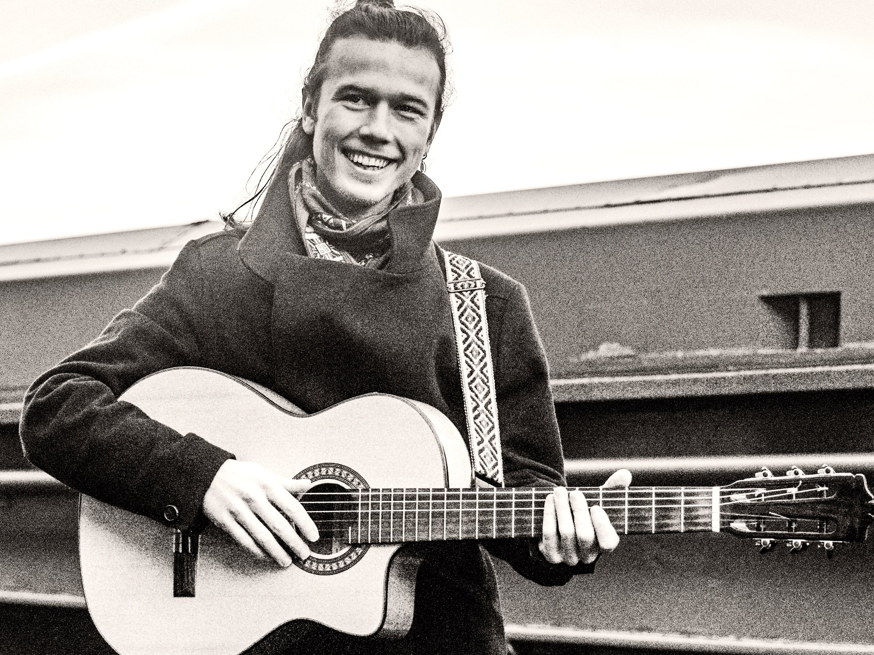 Erik Sjöholm smiling, playing an acoustic guitar outdoors by the water
